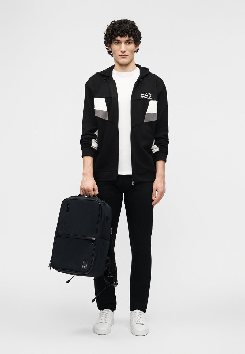 Giovane uomo con giacca e pantaloni bianco e nero tiene uno zaino nero con scarpe da ginnastica bianche davanti a uno sfondo bianco semplice.