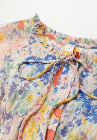 Blouse fleurie avec col volanté, devant boutonné et cordon noué, présentant des fleurs multicolores en bleu, jaune, vert et orange.