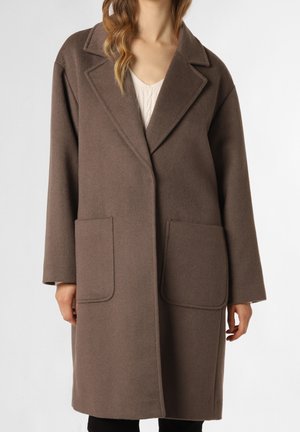 Femme portant un long manteau en laine marron avec de larges revers, deux grandes poches avant, et un haut en tricot crème en dessous.