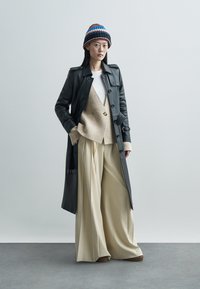 Black leather trench coat, beige sweater, white shirt, and beige wide-leg trousers. Black and multi-colored knit hat. Smooth textures.