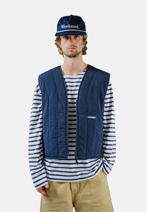 Mężczyzna w granatowej czapce z napisem „Weekend.”, granatowej pikowanej kamizelce na białej koszuli z długim rękawem w granatowe paski i beżowych spodniach, stojący na jednolitym tle.