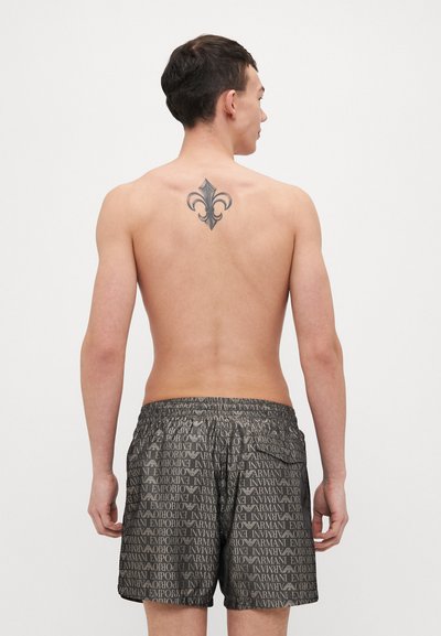 Črne kopalne hlače z ponovljenim vzorcem besedila "Emporio Armani", elastičnim pasom in gladko teksturo; nosi jih moški model, ki je prikazan od zadaj.