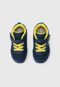 Marineblå nett-sneakers med gule lisser, borrelåsstropp og hvit såle. Har en lyseblå detalj nær hælen og polstret innside.