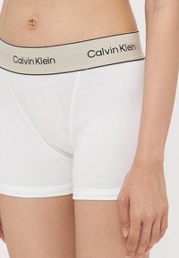 Bijele pamučne bokserice s širokim bež pojasom na kojem se nalazi crni tekst "Calvin Klein". Mekana tekstura i prilagođeni dizajn.