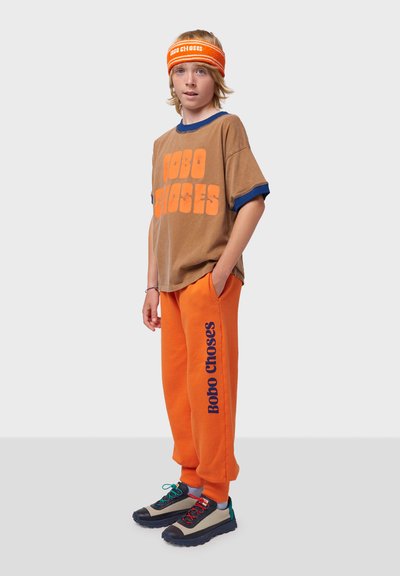 Braunes T-Shirt mit kurzen Ärmeln und orangefarbenem "Bobo Choses"-Druck, orangefarbene Sweatpants mit blauem Druck und mehrfarbige Sneakers. Strukturiertes Stirnband.