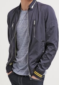 Giacca bomber blu navy con una texture liscia, dotata di cerniere argentate, tasche laterali e polsini a righe gialle, indossata sopra una maglietta grigia.