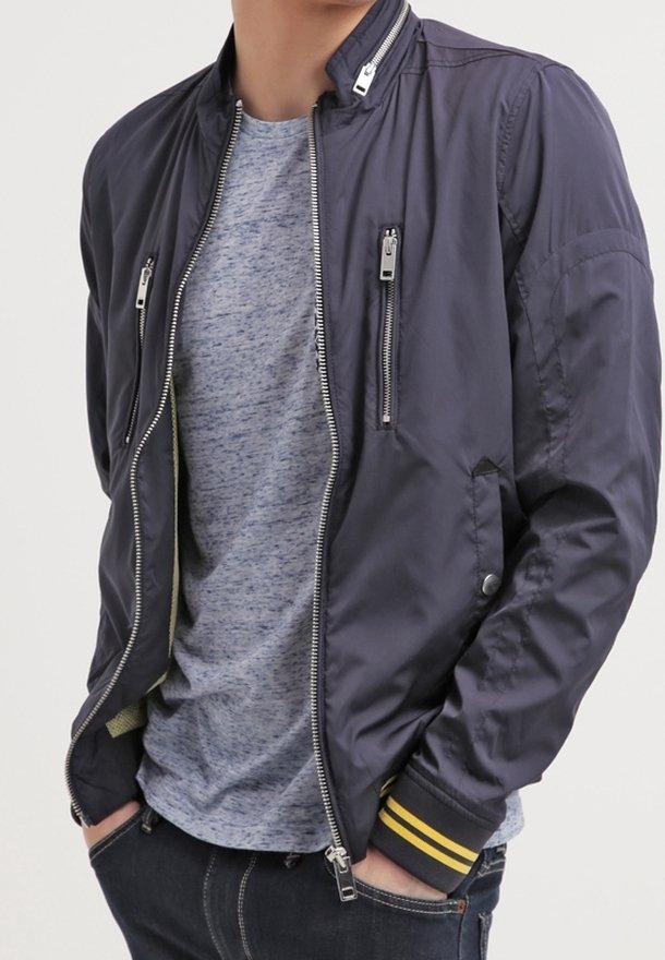 Giacca bomber blu navy con una texture liscia, dotata di cerniere argentate, tasche laterali e polsini a righe gialle, indossata sopra una maglietta grigia.