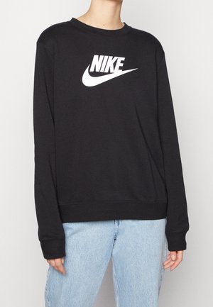 Osoba ubrana w czarną bluzę Nike z białym logo i jasnoniebieskie dżinsy, stojąca na tle jednolitego białego tła.