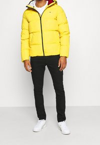 Žlutá péřová bunda s předním zipem, kapucí a černými akcenty. Nosit s černými kalhotami a bílými teniskami, které mají hladkou texturu.