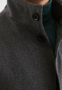 Grauer strukturierter Mantel mit hohem Kragen, versehen mit zwei schwarzen Knöpfen. Trägt man über einem dunkelgrünen Rollkragenpullover.