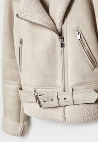 Veste beige en fausse shearling avec fermeture éclair argentée, deux poches zippées et ceinture assortie avec boucle métallique et œillets.