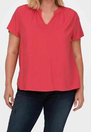 Kobieta w czerwonej krótkorękawowej bluzce z dekoltem w kształcie litery V oraz ciemnoniebieskich dżinsach, stojąca na tle jednolitego białego tła.