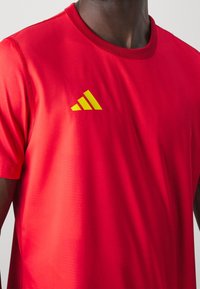 Röd idrottströja i slät tyg, med rund hals och korta ärmar. Har en gul Adidas-logo på vänster bröst.