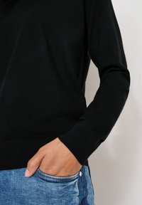 Maglione nero lavorato a maglia con collo rotondo e polsini a coste, indossato sopra un paio di jeans blu. La mano riposa nella tasca, mostrando i dettagli della trama.