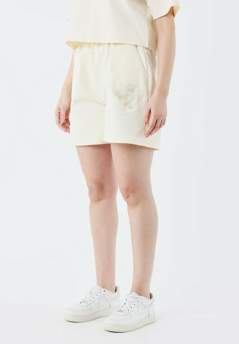 Shorts couleur crème en tissu doux, arborant un motif de cœur sur la jambe gauche. Les shorts ont une coupe décontractée et des poches latérales.