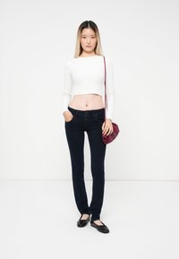 Jeans ajustados azul oscuro, top blanco de manga larga y corta, zapatos planos negros y un pequeño bolso de hombro burdeos. Fondo sencillo.