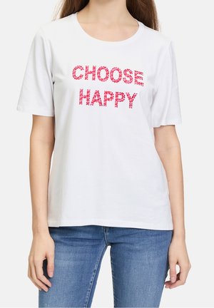 Femme portant un T-shirt blanc à manches courtes avec l'inscription "CHOOSE HAPPY" en lettres florales roses, associée à un jean bleu.