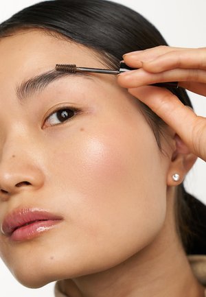 Applicatore a pennello tenuto vicino all'eyebrow, con una pelle liscia e neutra, un leggero fard e labbra lucide. Il riflesso della luce nell'occhio mette in risalto i dettagli.