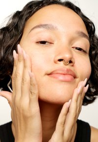 Person med lockigt mörkt hår som rör vid sitt ansikte. De har en gyllene septumring och ljusrosa, långt manikyrerade naglar. Snygg hudstruktur.