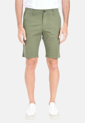 Uomo che indossa pantaloncini verde oliva fino al ginocchio, scarpe da ginnastica bianche e una camicia bianca su uno sfondo chiaro e uniforme.