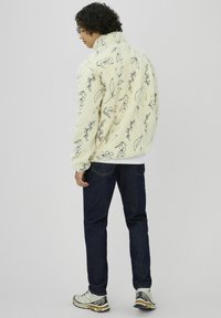Veste en polaire crème avec motif botanique noir, dotée d'un col montant et d'une coupe décontractée. Associée à un jean denim bleu foncé et des baskets.
