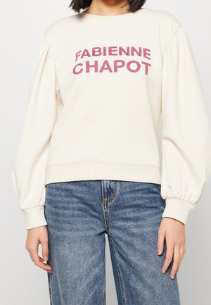Femme portant un sweat crème à manches bouffantes avec un texte rose "FABIENNE CHAPOT", associé à un jean bleu en denim.