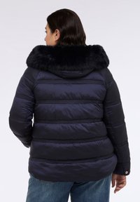 Marineblaue Pufferjacke mit horizontalen Nähten, Kapuze aus Kunstfell und glattem Stoff. Verfügt über versteckte Taschen und strapazierfähige Ärmel. Lässige Passform.
