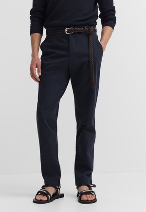 Mann trägt marineblaue Hose mit geflochtenem braunem Gürtel, schwarze Sandalen und einen dunklen Langarmpullover, steht vor einem einfarbigen Hintergrund.