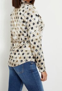 Blouse en soie avec un fond beige orné de motifs circulaires noirs et dorés, manches longues et ourlet arrondi, associée à un jean bleu.