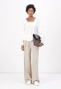 Witte blouse met lange mouwen en groene accenten, relaxed fit, geplooide halslijn; gecombineerd met beige wijde broek en zwarte clutch.
