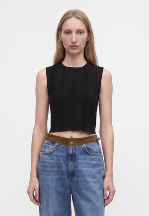 Jeune femme aux longs cheveux blonds portant un crop top noir sans manches et un jean bleu taille haute avec une ceinture marron sur un fond uni.