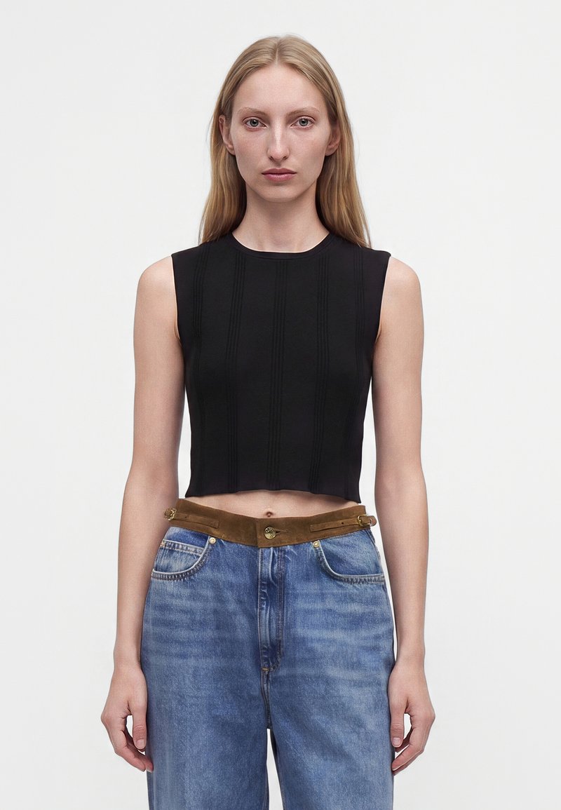 Jeune femme aux longs cheveux blonds portant un crop top noir sans manches et un jean bleu taille haute avec une ceinture marron sur un fond uni.