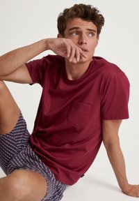 Bordeauxrotes T-Shirt mit kurzen Ärmeln und Tasche, kombiniert mit gemusterten Shorts in Blau- und Rottönen. Glatter Stoff und lässige Passform.