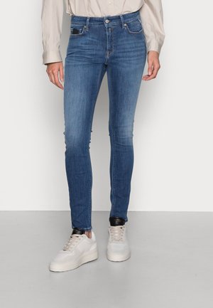 Persoon die een nauwsluitende blauwe spijkerbroek en witte sneakers draagt en tegen een effen achtergrond staat.