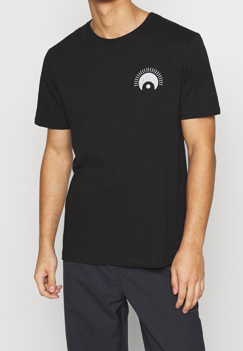 Homme portant un T-shirt noir à manches courtes avec un petit motif blanc en demi-cercle représentant des rayons de soleil sur le côté gauche de la poitrine et un pantalon foncé.