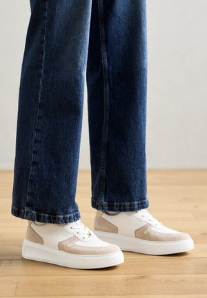 Persoon draagt witte en beige sneakers gecombineerd met donkerblauwe wijde jeans, staand op een houten vloer.