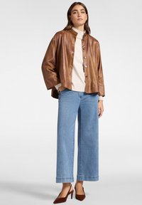 Veste en cuir marron avec un col arrondi et des boutons, portée sur un pull léger et associée à un jean en denim bleu à jambes larges et des talons bruns.