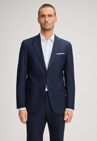 Abito su misura blu navy con chiusura a due bottoni, bavero a lancia e tasche laterali, abbinato a una camicia da uomo leggera con motivo.