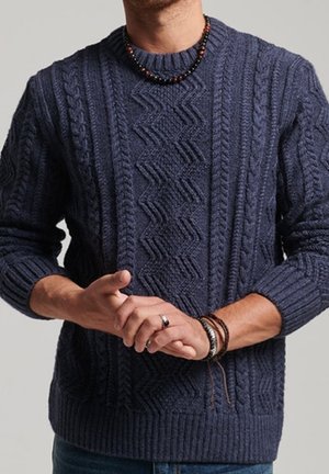 Mann trägt einen dunkelblauen Zopfpullover, Perlenkette, mehrere Armbänder und einen Ring, mit vorgefassten Händen.