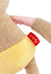 Plush toy with a textured tan body, yellow base, and pink checkered fabric arms. Features a red brand tag on the side.