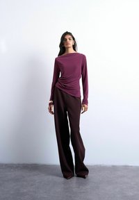 Femme portant un haut violet à manches longues avec un côté froncé, associé à un pantalon large foncé et des chaussures marron à bout pointu, debout sur un sol gris.