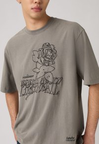 Άνδρας που φοράει γκρι μπλουζάκι με μαύρο περίγραμμα τριαντάφυλλου και τη λέξη "Permanent" στο μπροστινό μέρος, λογότυπο Levi's κοντά στο τελείωμα.