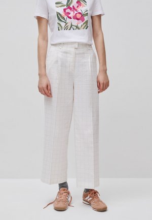 Person trägt weiße, weit geschnittene Hosen mit dezentem Karomuster, weißes T-Shirt mit Blumenmuster, graue Socken und hellbraune Sneakers mit offenen Schnürsenkeln.