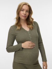 Terhes nő, aki világos szőke haj�ú, olíva színű, bordázott hosszú ujjú toppot visel, és óvatosan a hasát simogatja az egyik kezével.