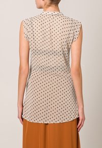 Blusa sin mangas beige con un patrón transparente de puntos en gris oscuro. Dobladillo redondeado y un sutil detalle de collar. Tejido suave y ligero.