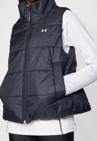 Marineblaue Steppweste mit hohem Kragen, Reißverschluss und genähten Abschnitten. Mit einem Logo auf der Brust und seitlichen Taschen. Glattes, leichtes Material.