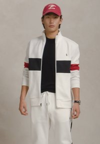 Νεαρός άνδρας με λευκή ζακέτα με φερμουάρ και μαύρες και κόκκινες ρίγες, λευκά παντελόνια, μαύρο πουκάμισο, κόκκινο καπέλο μπέιζμπολ και μαύρο ρολόι χειρός.