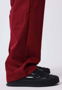 Sneakers nere con suola testurizzata e dettagli in argento, abbinate a pantaloni larghi color rosso intenso in tessuto liscio, che coprono parzialmente le calzature.