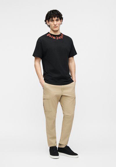 Egy fiatal férfi göndör hajjal, zsebeibe dugott kézzel áll, fekete pólót visel narancssárga felirattal, bézs cargo nadrágot és fekete sportcipőt.