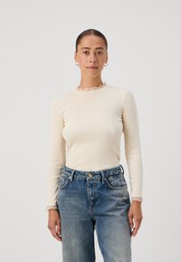 Long-sleeve cream ribbed top with a ruffled neckline, paired with high-waisted faded blue jeans. Simple design, soft texture.
